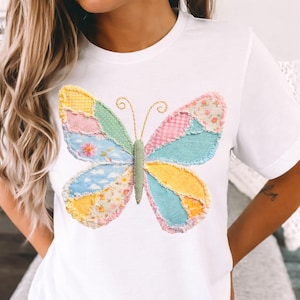 May include: White t-shirt featuring a colorful butterfly design. The butterfly is made of patchwork fabric in various pastel colors, including pink, blue, yellow, and green. The butterfly has a green body and gold wire antennae. The shirt is a casual, everyday wear item.