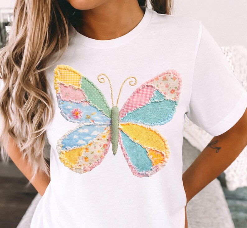 May include: White t-shirt featuring a colorful butterfly design. The butterfly is made of patchwork fabric in various pastel colors, including pink, blue, yellow, and green. The butterfly has a green body and gold wire antennae. The shirt is a casual, everyday wear item.
