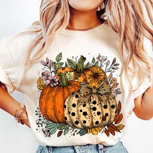 May include: Cream-colored sweatshirt featuring a colorful fall-themed graphic. The design includes three pumpkins in shades of orange and yellow, adorned with a bow and surrounded by floral and leaf accents. A seasonal apparel item.