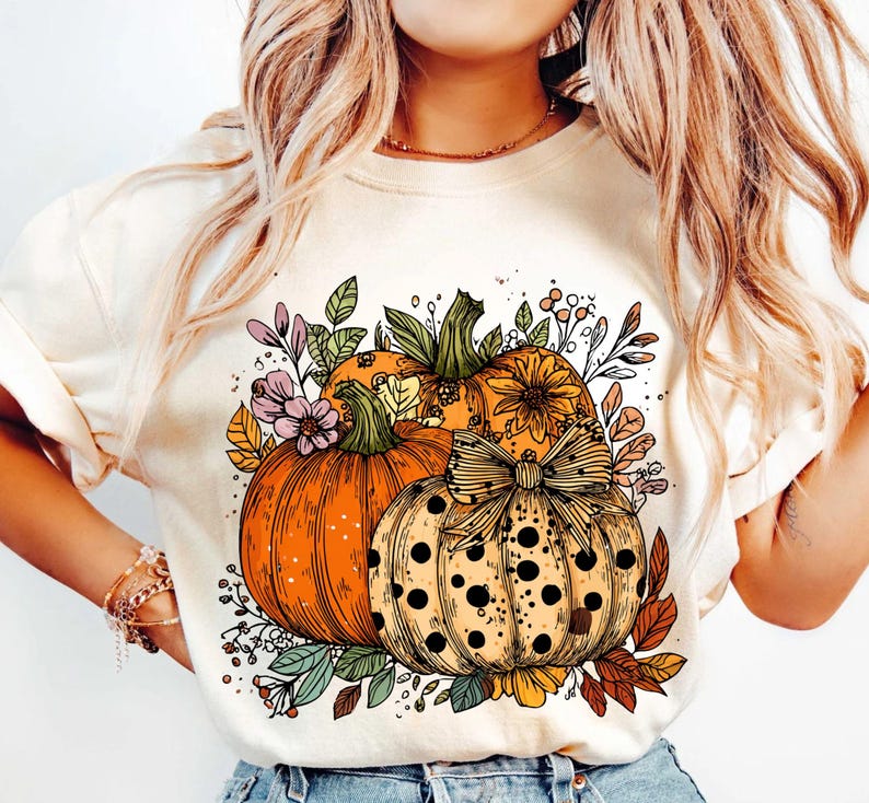 May include: Cream-colored sweatshirt featuring a colorful fall-themed graphic. The design includes three pumpkins in shades of orange and yellow, adorned with a bow and surrounded by floral and leaf accents. A seasonal apparel item.