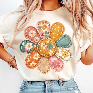 Może przedstawiać: Kremowy t-shirt z dużym, kolorowym kwiatowym wzorem. Kwiat ma płatki w stylu patchwork w r&oacute;żnych kolorach i wzorach, w tym nadruki kwiatowe i kropki. Projekt jest wyśrodkowany na koszulce, tworząc żywy i przyciągający wzrok wygląd.