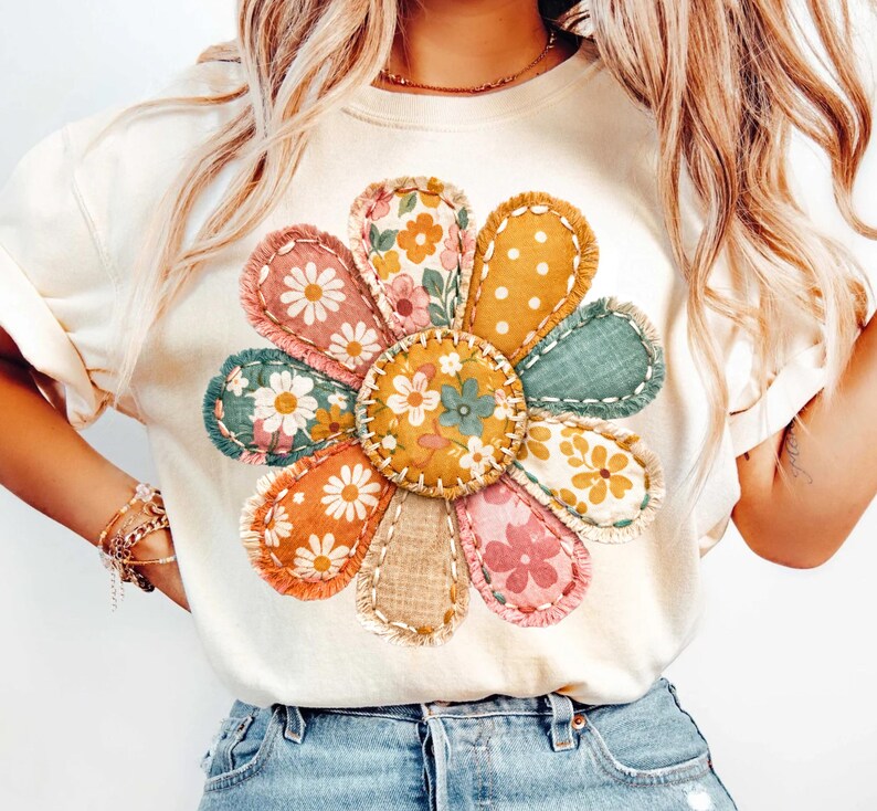Może przedstawiać: Kremowy t-shirt z dużym, kolorowym kwiatowym wzorem. Kwiat ma płatki w stylu patchwork w r&oacute;żnych kolorach i wzorach, w tym nadruki kwiatowe i kropki. Projekt jest wyśrodkowany na koszulce, tworząc żywy i przyciągający wzrok wygląd.