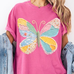 May include: A pink t-shirt featuring a colorful patchwork butterfly design. The butterfly has wings in shades of yellow, blue, pink, and green, with a textured, stitched appearance. The shirt has short sleeves and a relaxed fit.