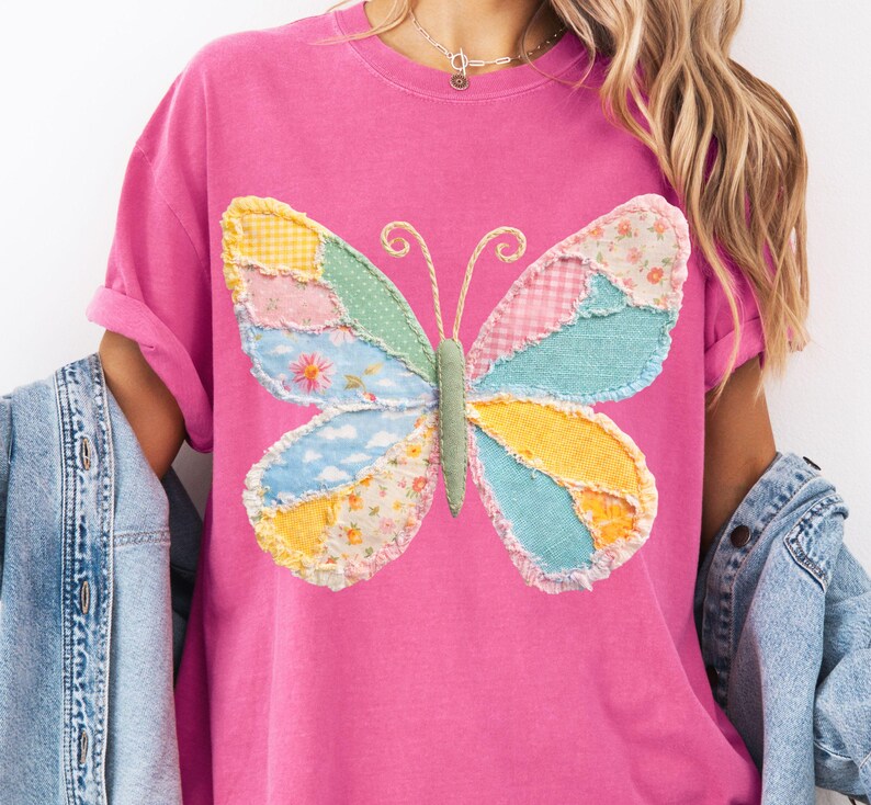 May include: A pink t-shirt featuring a colorful patchwork butterfly design. The butterfly has wings in shades of yellow, blue, pink, and green, with a textured, stitched appearance. The shirt has short sleeves and a relaxed fit.