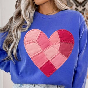 Puede incluir: Sudadera azul con un diseño de corazón de patchwork en tonos rosas y rojos. El corazón está hecho de cuadrados de tela cosidos, dando una apariencia texturizada. La sudadera tiene mangas largas y cuello redondo. El diseño está centrado.