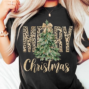 K&ouml;nnte beinhalten: Schwarzes T-Shirt mit einem Weihnachtsdesign. Das Wort "MERRY" ist im Leopardenmuster gehalten, und das Wort "Christmas" ist in goldfarbenem Glitzer geschrieben. Ein gr&uuml;ner Weihnachtsbaum mit goldener Schleife befindet sich in der Mitte.