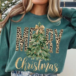 K&ouml;nnte beinhalten: T&uuml;rkisgr&uuml;nes Sweatshirt mit den Worten "MERRY Christmas" in goldglitzernder Schrift. Das Wort "MERRY" hat ein Leopardenmuster. Ein gr&uuml;ner Weihnachtsbaum mit goldener Schleife und Lichtern befindet sich in der Mitte.