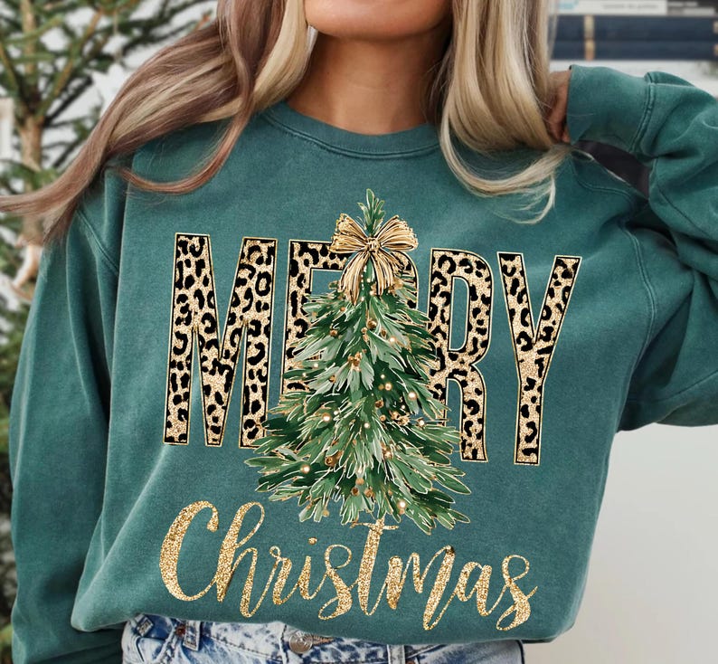 K&ouml;nnte beinhalten: T&uuml;rkisgr&uuml;nes Sweatshirt mit den Worten "MERRY Christmas" in goldglitzernder Schrift. Das Wort "MERRY" hat ein Leopardenmuster. Ein gr&uuml;ner Weihnachtsbaum mit goldener Schleife und Lichtern befindet sich in der Mitte.