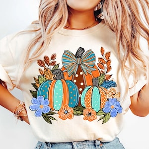 May include: Cream-colored sweatshirt featuring a fall-themed graphic. The design includes three pumpkins in shades of orange and teal, adorned with polka dots and a large bow. Floral accents in blue and orange complete the autumn-inspired look.