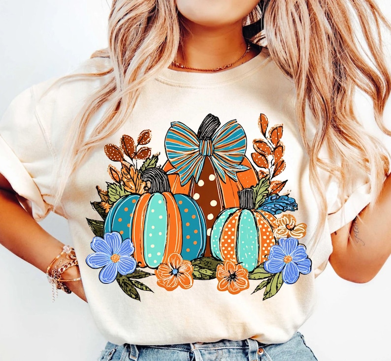 May include: Cream-colored sweatshirt featuring a fall-themed graphic. The design includes three pumpkins in shades of orange and teal, adorned with polka dots and a large bow. Floral accents in blue and orange complete the autumn-inspired look.