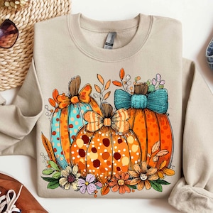 May include: A beige sweatshirt featuring a colorful graphic of three pumpkins. The pumpkins are orange with polka dots and bows, surrounded by flowers and leaves. The design has a fall theme.