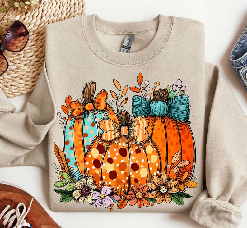 May include: A beige sweatshirt featuring a colorful graphic of three pumpkins. The pumpkins are orange with polka dots and bows, surrounded by flowers and leaves. The design has a fall theme.
