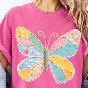 May include: Pink t-shirt featuring a colorful patchwork butterfly design. The butterfly wings are made of various patterned fabrics in shades of yellow, pink, blue, and green. The shirt has a crew neck and short sleeves.