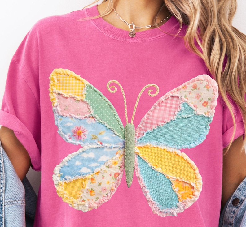 May include: Pink t-shirt featuring a colorful patchwork butterfly design. The butterfly wings are made of various patterned fabrics in shades of yellow, pink, blue, and green. The shirt has a crew neck and short sleeves.
