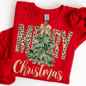 May include: A red sweatshirt with the words "MERRY Christmas" in a gold glitter script. The word "MERRY" has a leopard print pattern. A green Christmas tree with gold ornaments and a gold bow is in the center.