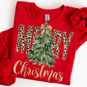 K&ouml;nnte beinhalten: Ein rotes Sweatshirt mit dem Schriftzug "MERRY Christmas" in goldfarbenem Glitzer. Das Wort "MERRY" hat ein Leopardenmuster. Ein gr&uuml;ner Weihnachtsbaum mit goldenen Ornamenten und einer goldenen Schleife befindet sich in der Mitte.