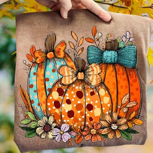 May include: A brown t-shirt with a vibrant autumn design. The design showcases three pumpkins adorned with polka dots and bows, complemented by flowers and leaves. The pumpkins are orange, teal, and brown.