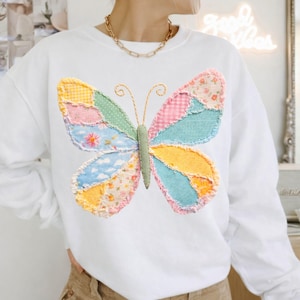 May include: White sweatshirt featuring a large, colorful butterfly applique. The butterfly design incorporates various fabric patterns in shades of yellow, pink, blue, and green. The sweatshirt has long sleeves and a crew neck.