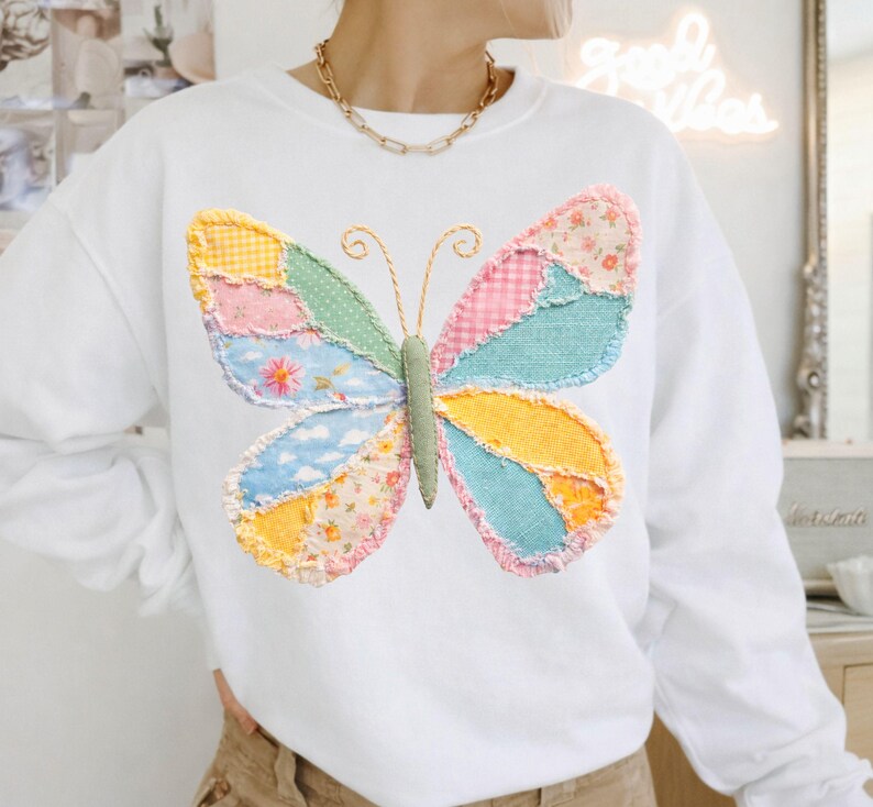 May include: White sweatshirt featuring a large, colorful butterfly applique. The butterfly design incorporates various fabric patterns in shades of yellow, pink, blue, and green. The sweatshirt has long sleeves and a crew neck.