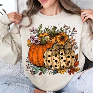 May include: Cream-colored sweatshirt featuring a colorful illustration of pumpkins and fall foliage. The design includes three pumpkins in shades of orange and yellow, accented with leaves and flowers, perfect for autumn.