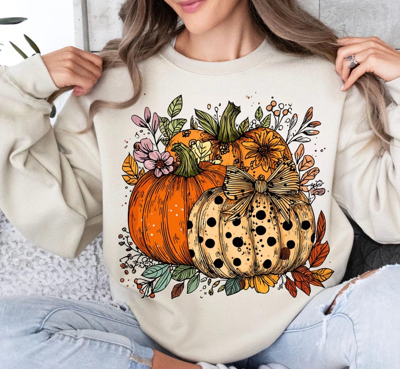 May include: Cream-colored sweatshirt featuring a colorful illustration of pumpkins and fall foliage. The design includes three pumpkins in shades of orange and yellow, accented with leaves and flowers, perfect for autumn.