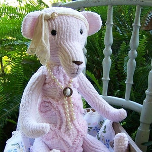 Puede incluir: Un oso de peluche rosa claro con una diadema color crema y un collar de perlas. El oso está sentado en una caja de madera con una tela estampada floral. El oso tiene ojos negros y nariz marrón.