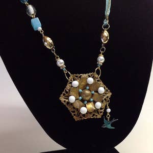 May include: A gold necklace with a large flower pendant featuring white and turquoise beads. The necklace is made with a variety of beads, including gold, white, and turquoise.
