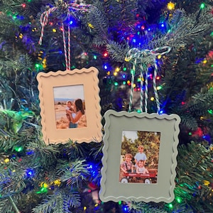 Puede incluir: Dos adornos navideños con forma de marco de fotos con bordes festoneados colgados de un árbol de Navidad. Uno es de color beige, el otro verde salvia. Cada uno contiene una fotografía en color y está suspendido por hilo.