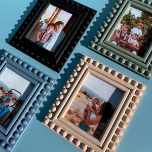 Puede incluir: Cuatro marcos de fotos decorativos en negro, gris y blanquecino, cada uno con un borde de cuentas. Los marcos contienen fotografías de personas. Los marcos están dispuestos sobre una superficie azul claro, proyectando sombras.