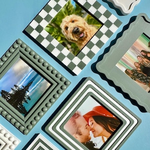 May include: An assortment of decorative picture frames in various styles and colors. The frames hold photographs of a dog, a landscape, and a couple. The frames are in shades of green, white, and gray, with different patterns and textures. The background is a solid light blue.