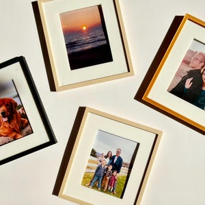 Puede incluir: Cuatro marcos de fotos rectangulares con paspartú blanco, cada uno con una fotografía diferente. Los marcos son negros, de madera clara y dorados, y muestran imágenes de una puesta de sol, un perro dorado, una pareja y una familia de cinco.