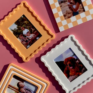 May include: Four decorative picture frames in various colors, including orange, white, and a checkered pattern. Each frame holds a photograph. The frames have a scalloped edge design and are arranged on a pink surface.