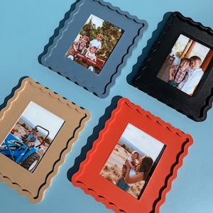 Può includere: Quattro cornici colorate con bordi smerlati. Le cornici sono blu, nere, beige e rosse. Ogni cornice contiene una foto di persone. Le cornici sono disposte su una superficie azzurra.