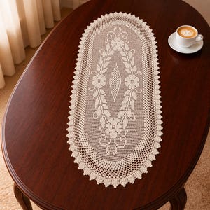 Op de afbeelding: Een crèmekleurige, ovale kanten tafelloper met een bloemen- en bladerenpatroon. De loper ligt op een donkerbruine houten tafel. Een witte koffiekop met een latte staat op de tafel.