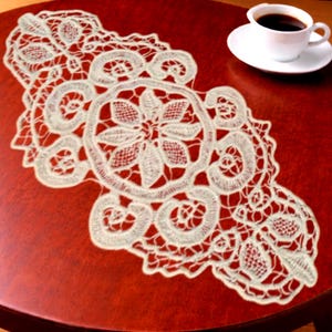 May include: An off-white lace table runner featuring an intricate openwork design with central floral motifs and swirling scroll patterns. The delicate textile is elongated with scalloped edges, showcasing detailed needlework and fine thread construction.
