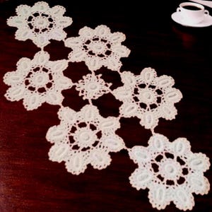 May include: A white crocheted table runner featuring a series of interconnected floral motifs with intricate openwork patterns. The lace-like textile is composed of several circular, scalloped-edge segments joined together in a linear arrangement.