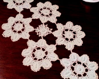 May include: A white crocheted table runner featuring a series of interconnected floral motifs with intricate openwork patterns. The lace-like textile is composed of several circular, scalloped-edge segments joined together in a linear arrangement.