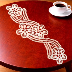 Puede incluir: Un camino de mesa de encaje blanco con un patrón floral repetitivo de pétalos interconectados y detalles calados intrincados, dispuesto en un diseño fluido y diagonal sobre una mesa redonda de madera oscura pulida.