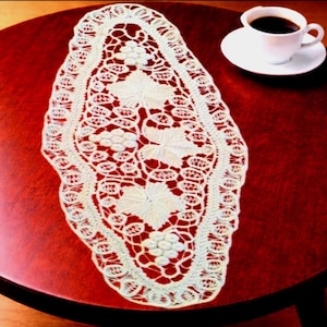 May include: An oval, cream-coloured lace doily with a floral pattern, featuring grape clusters and leaves, sits on a dark red wooden table. A white teacup and saucer with coffee are to the right, creating a cosy scene.