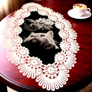 May include: An oval-shaped black fabric table runner featuring a white crocheted lace border. The center of the runner displays a detailed monochromatic graphic print of a cat looking upward, positioned below a cluster of white flowers.