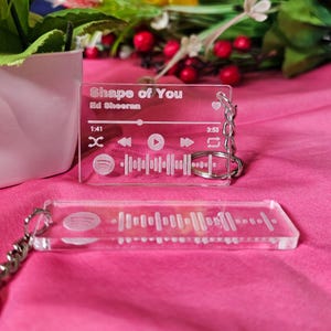Könnte beinhalten: Transparenter Acryl-Schlüsselanhänger mit den Songdetails von "Shape of You" von Ed Sheeran. Das Design umfasst Songtitel, Künstler, Wiedergabesteuerung und einen Spotify-Code. Ein zweiter Schlüsselanhänger ist ebenfalls sichtbar.