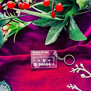 Puede incluir: Un llavero acrílico transparente con el texto "Shape of You" de Ed Sheeran. El llavero presenta un diseño estilo Spotify con un botón de reproducción, una barra de progreso de la canción y un código QR. Está unido a un llavero plateado. El fondo es una tela roja oscura con vegetación.
