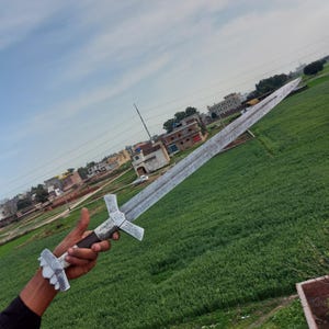May include: A long, silver sword with a detailed blade and ornate cross-shaped hilt. The handle is dark brown. The sword is held against a backdrop of a green field and a cloudy sky.