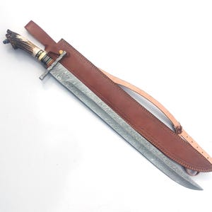 May include: An ornate sword with a patterned blade and a brown leather sheath. The handle has a decorative design and a light-colored grip. The sheath has a strap.