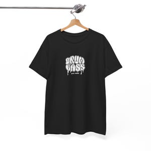 Puede incluir: Camiseta negra con la inscripción blanca "DRUM & BASS" en una fuente estilizada. La camiseta está colgada de una percha de madera sobre un fondo blanco. El texto "NOISE UNDER" está debajo.