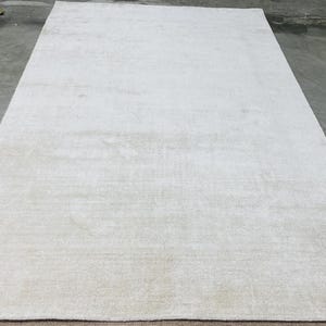 May include: A large, rectangular, off-white rug. The rug has a soft, textured appearance and is placed on a concrete floor. The neutral color and simple design suggest a modern or minimalist style, suitable for various interior settings.