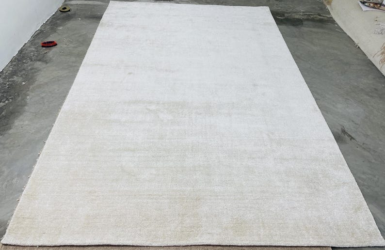 May include: A large, rectangular, off-white rug. The rug has a soft, textured appearance and is placed on a concrete floor. The neutral color and simple design suggest a modern or minimalist style, suitable for various interior settings.
