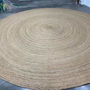 K&ouml;nnte beinhalten: Ein gro&szlig;er, runder Jute-Teppich in einem nat&uuml;rlichen, hellbraunen Farbton. Das Design des Teppichs zeigt konzentrische Kreise, die von der Mitte ausgehen und eine strukturierte Oberfl&auml;che bilden. Dieser Bodenbelag eignet sich f&uuml;r verschiedene Einrichtungsstile.