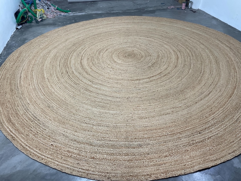 K&ouml;nnte beinhalten: Ein gro&szlig;er, runder Jute-Teppich in einem nat&uuml;rlichen, hellbraunen Farbton. Das Design des Teppichs zeigt konzentrische Kreise, die von der Mitte ausgehen und eine strukturierte Oberfl&auml;che bilden. Dieser Bodenbelag eignet sich f&uuml;r verschiedene Einrichtungsstile.