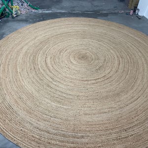 K&ouml;nnte beinhalten: Ein gro&szlig;er, runder Jute-Teppich mit einem konzentrischen Spiralmuster. Der Teppich hat eine nat&uuml;rliche, beige Farbe und scheint aus gewebten Fasern zu bestehen. Der Teppich liegt auf einem Betonboden.